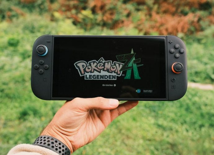 A hand holding a handheld gaming console displaying the Pokémon Legends game screen outdoors.