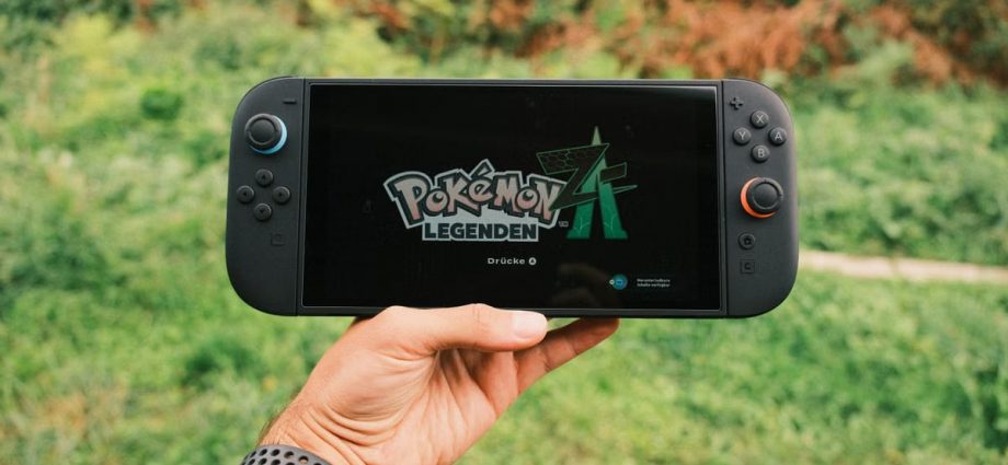 A hand holding a handheld gaming console displaying the Pokémon Legends game screen outdoors.