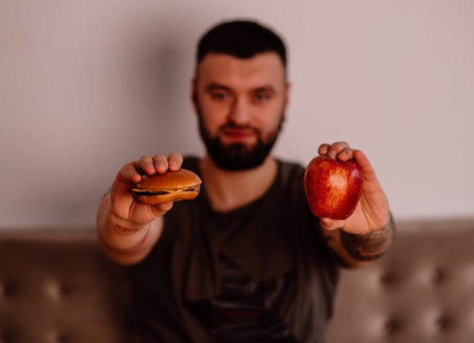 A man holds an apple and a cheeseburger, showcasing a choice between healthy and unhealthy food.