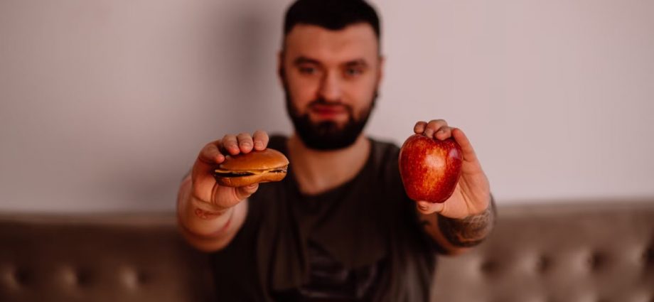 A man holds an apple and a cheeseburger, showcasing a choice between healthy and unhealthy food.